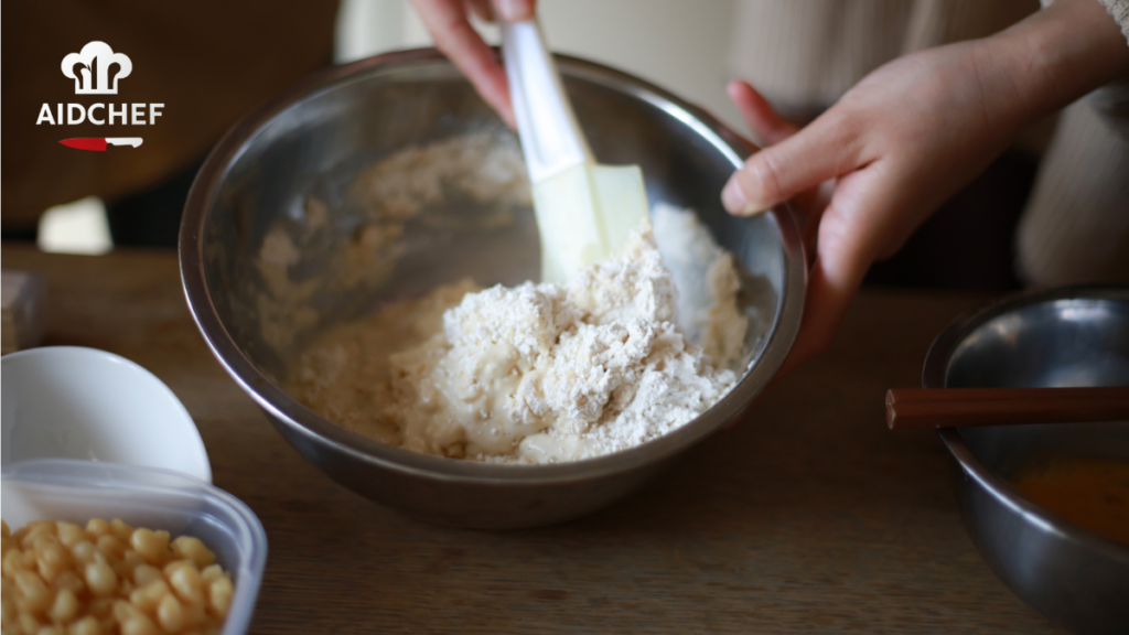 Espátula de silicona mezclando masa en bol de acero inoxidable sobre mesa de cocina